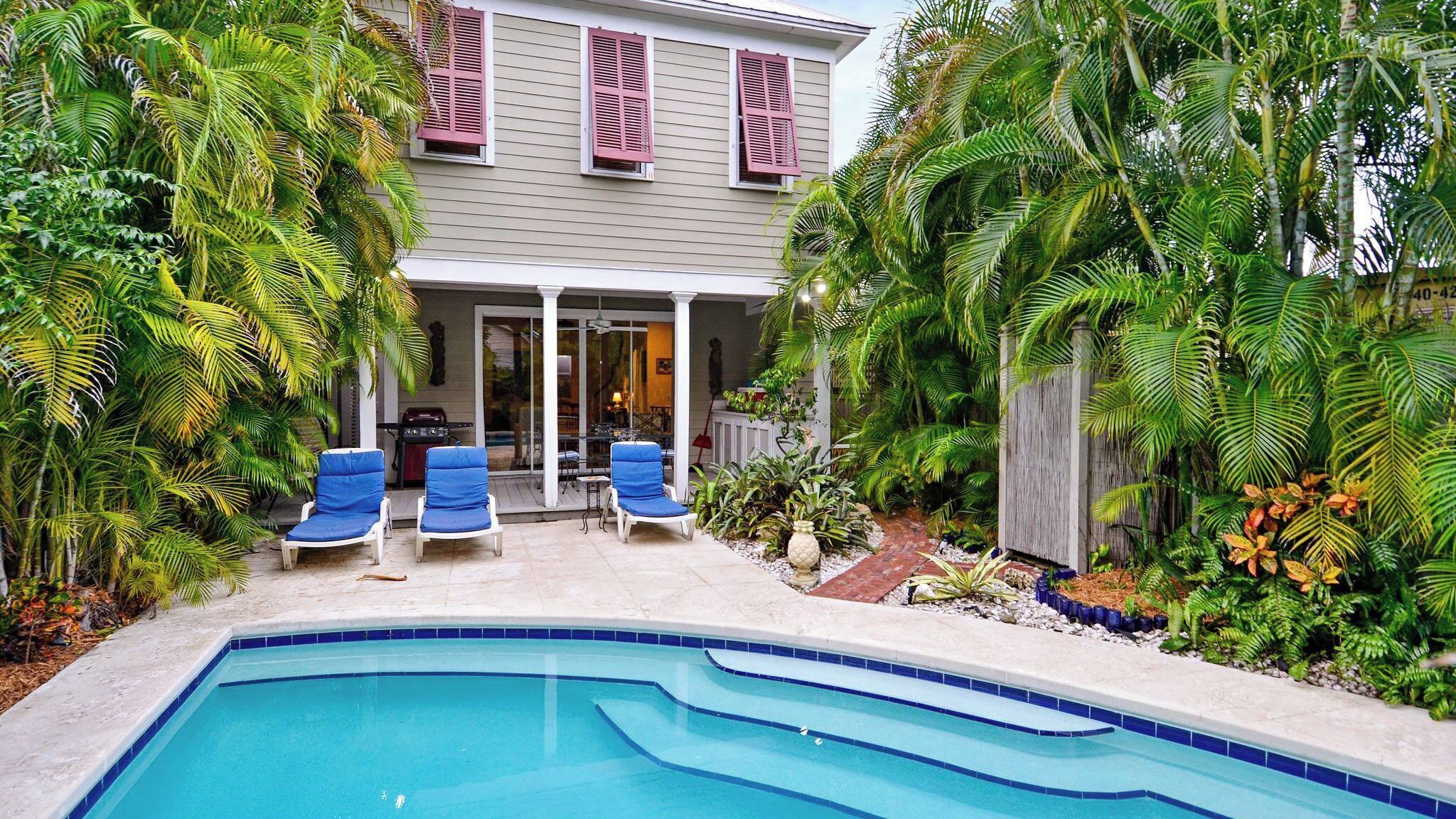 Ein Schwimmbad mit blauen Fliesen, umgeben von Palmen und einem Haus mit roten Fensterläden.