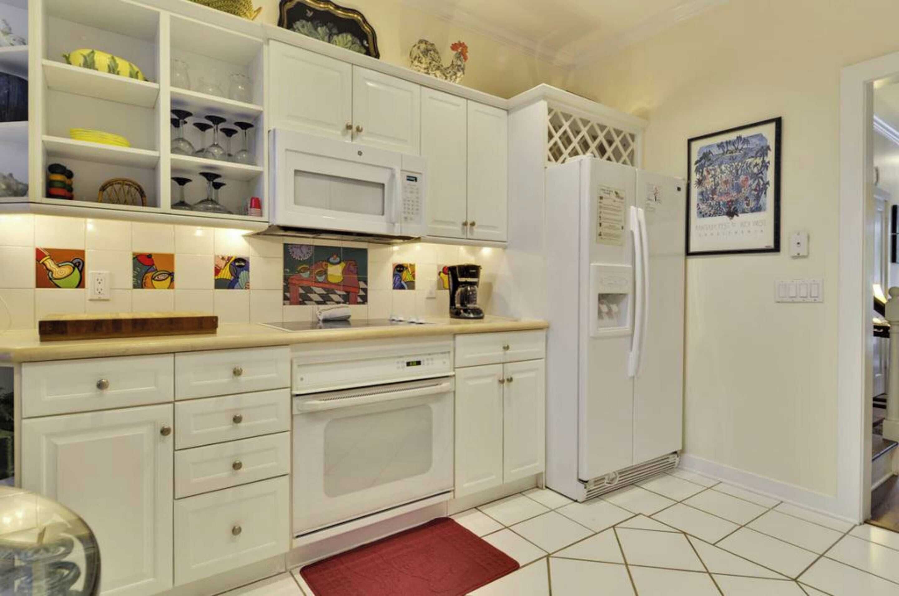 Una cucina con mobili bianchi, un forno a microonde, un frigorifero e uno schienale piastrellato con piastrelle colorate.