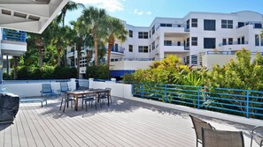 A rooftop deck with outdoor seating, palm trees, and a white building with balconies.
