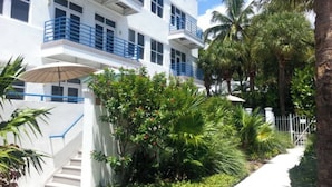 Un hôtel à plusieurs étages, avec des murs extérieurs blancs, des balustrades bleues et un jardin luxuriant agrémenté de palmiers et de plantes tropicales.