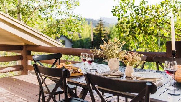 An outdoor dining area with wooden tables and chairs, surrounded by greenery and a wooden fence.