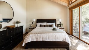 A bedroom with a large bed, wooden ceiling beams, a dresser, bedside tables, and a large window with a view of trees.