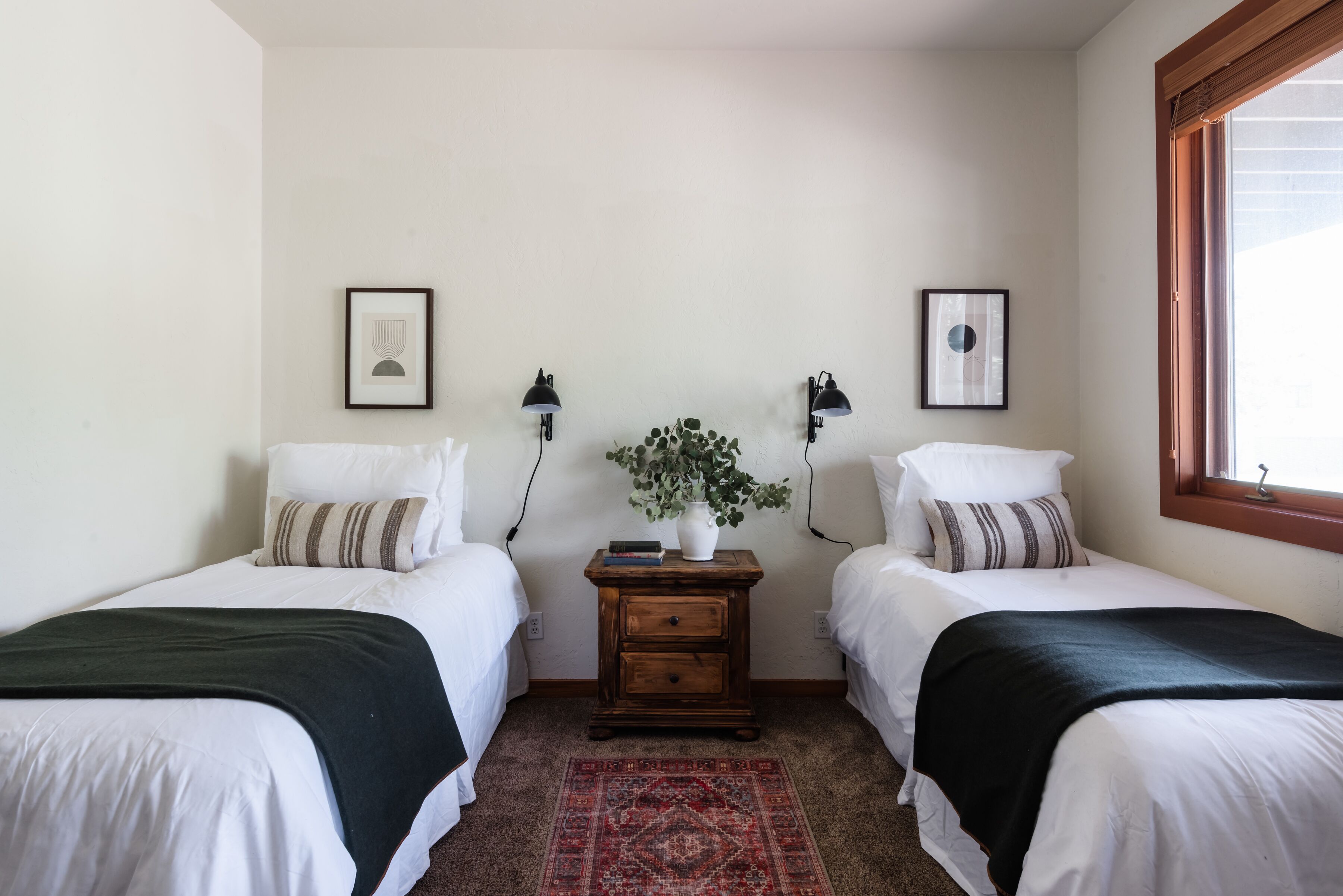 A room with two single beds, a nightstand with a plant, and framed pictures on the walls.