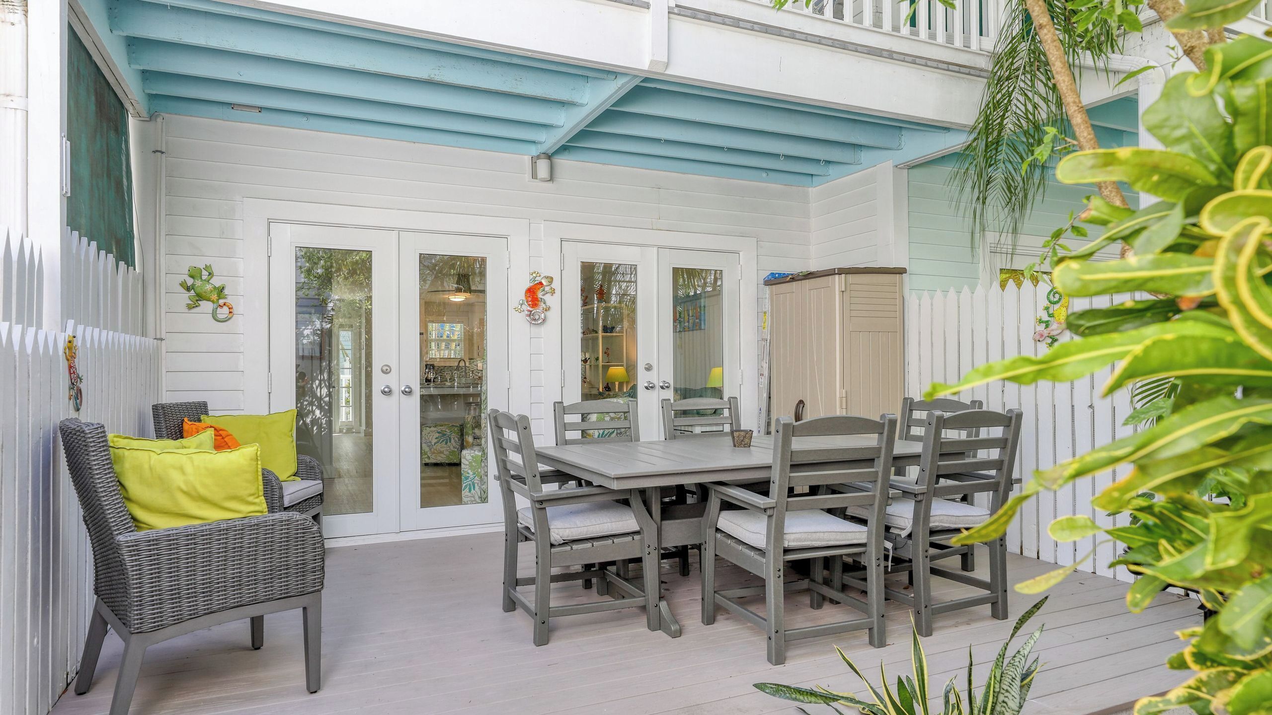 A covered patio with a table and chairs, a white picket fence, and a hanging plant.