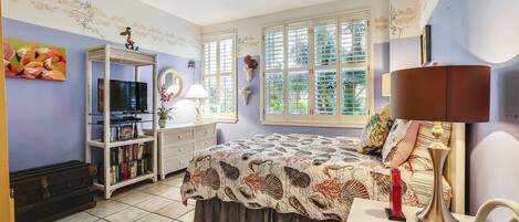 A bedroom with a bed, a TV, a bookshelf, and a window with shutters.