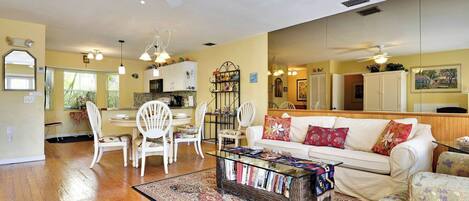A living room with a white sofa, a coffee table with books, a dining area with white chairs, and a kitchen in the background.