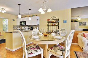 Uma sala de jantar com uma mesa redonda de vidro, cadeiras brancas com estofamento floral, uma cozinha com armários brancos e um micro-ondas, e um ventilador de teto.