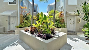 A residential area with a central planter with various plants, white houses with shutters, and a clear blue sky.