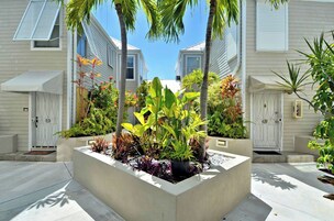 Zona residencial con un jardín central con diversas plantas, casas blancas con contraventanas y un cielo azul despejado.