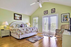 Un dormitorio con cama, mesitas de noche, una silla, ventilador de techo y dos ventanas con persianas.