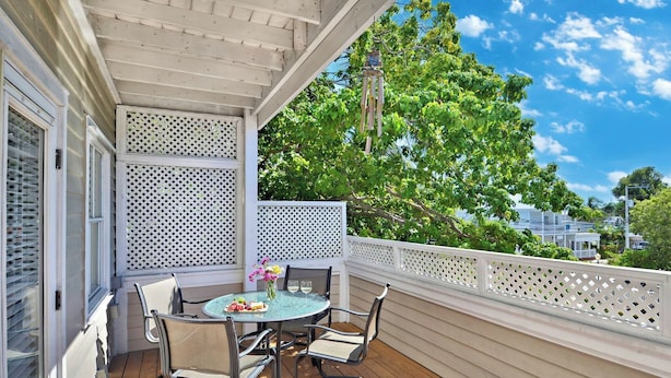 A covered patio with a table and chairs, a white lattice fence, and a view of trees and buildings.