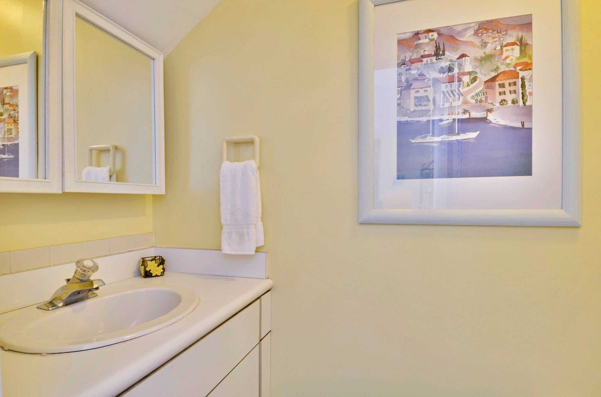 A bathroom with a sink, mirror, and a framed painting of a coastal scene.