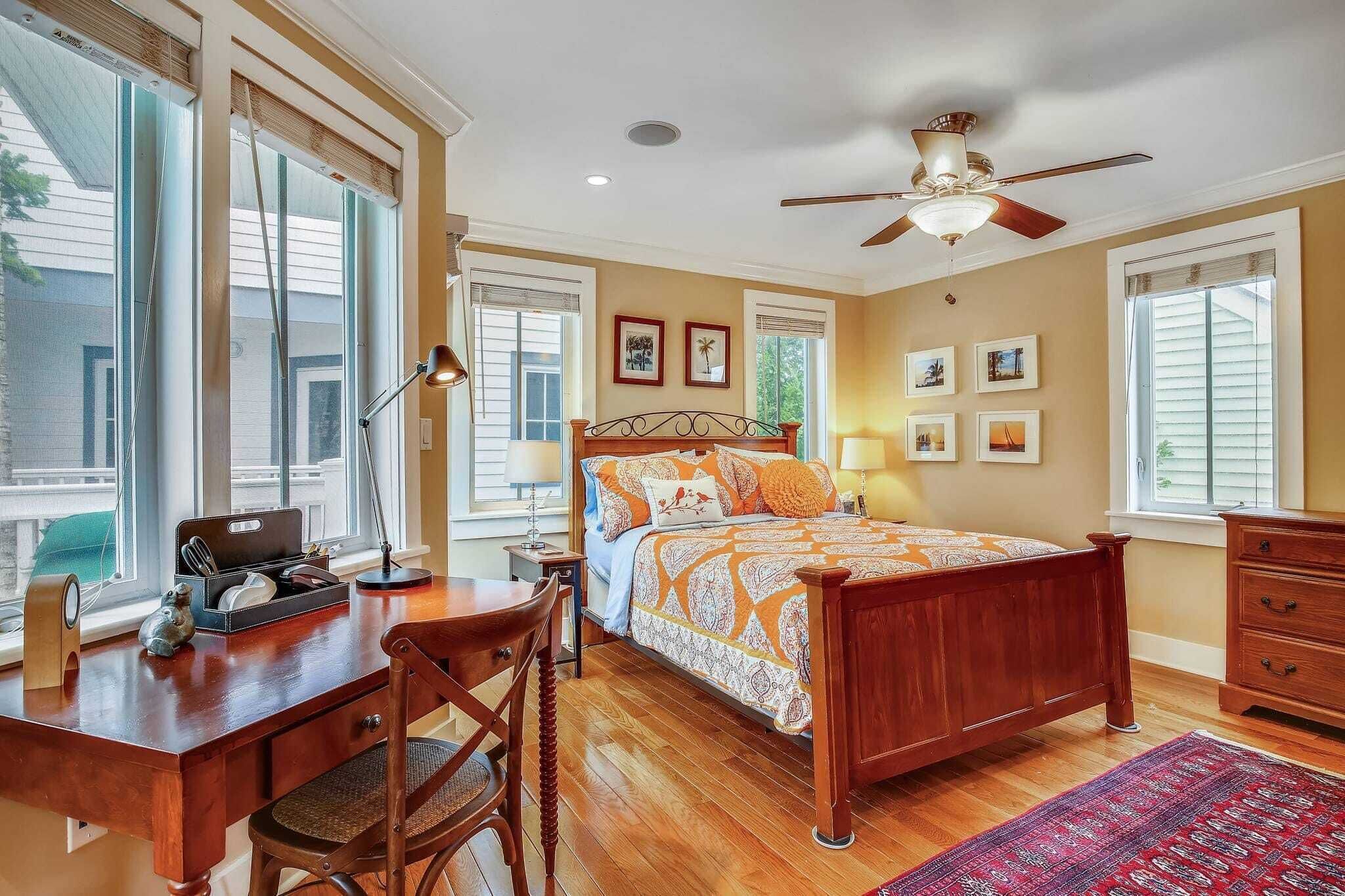 A bedroom with a wooden bed, a desk with a chair, a ceiling fan, and large windows.