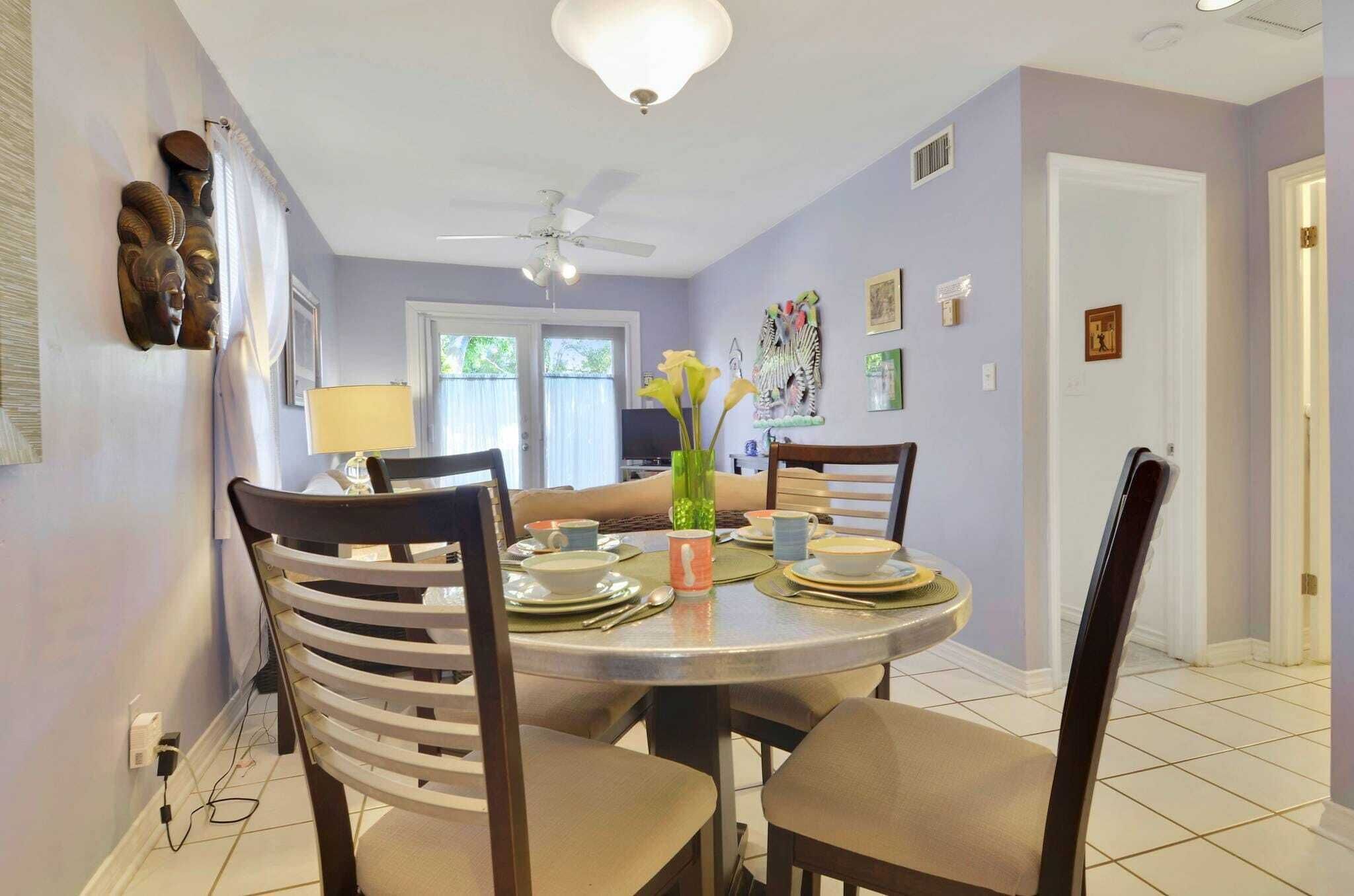 A dining area with a round table set for four, surrounded by chairs. There is a ceiling fan above the table, and the walls are painted in a light color.