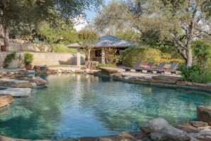 A clear swimming pool with a gazebo and lounge chairs, surrounded by rocks and trees.