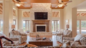 A spacious living room with a stone fireplace, two ceiling fans, and patterned furniture. - 10,000 Sq.Ft Lux Estate on 650 Acres with Amazing Pool, Spa & Hill Country Views (Marble Falls)