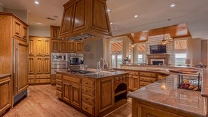A spacious kitchen with wooden cabinets, a central island, and a stone fireplace. - 10,000 Sq.Ft Lux Estate on 650 Acres with Amazing Pool, Spa & Hill Country Views (Marble Falls)