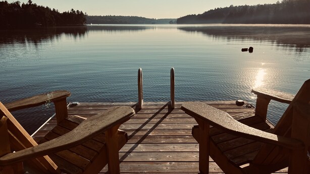 Eine Holzstege mit zwei Stühlen, die auf einen ruhigen See blicken, umgeben von Bäumen.