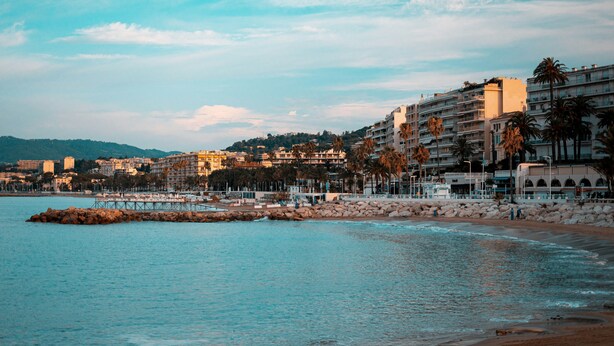 Un paisaje urbano costero con un rompeolas rocoso, edificios frente a la playa y palmeras.