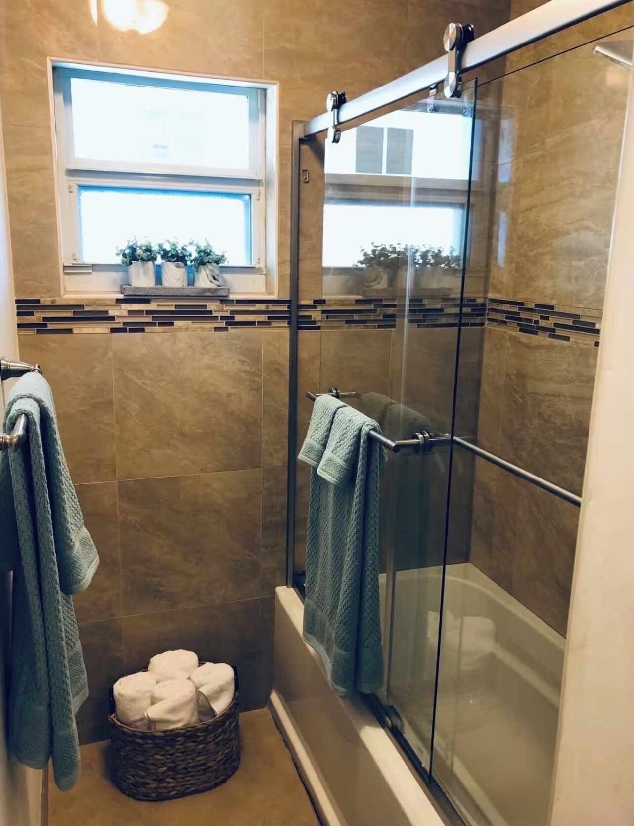 A modern bathroom with a glass-enclosed shower, a window with plants, and a basket of towels.