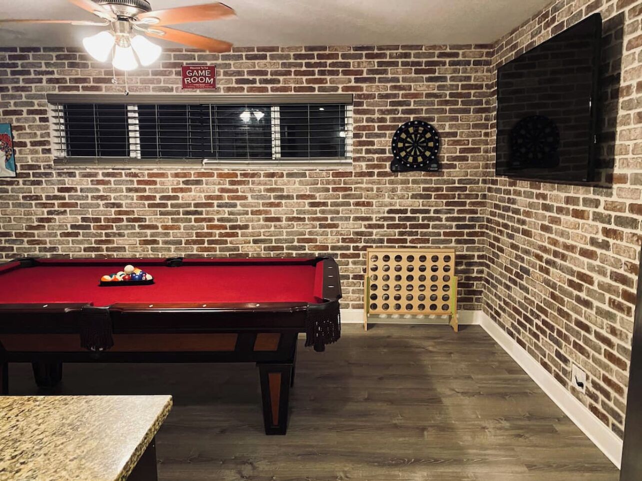 Une table de billard recouverte d’un drap de feutre rouge, un jeu de fléchettes et un ventilateur de plafond dans une pièce aux murs de briques et dotée d’une fenêtre.