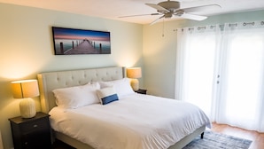 A neatly made bed with white bedding, a headboard, two bedside tables with lamps, a painting of a pier, and a view of the outdoors through a window with sheer curtains. - Skyline Breeze - Minutes from downtown Jensen Beach (Jensen Beach)