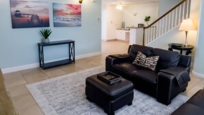 A living room with a black leather sofa, a coffee table, and a rug. There are two paintings on the wall, a staircase, and a kitchen in the background. - Skyline Breeze - Minutes from downtown Jensen Beach (Jensen Beach)
