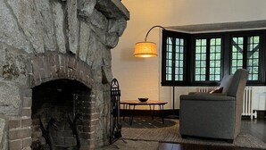 En stue med pejs af natursten, et bord, en stol og et vindue med udsigt over grøn bevoksning.