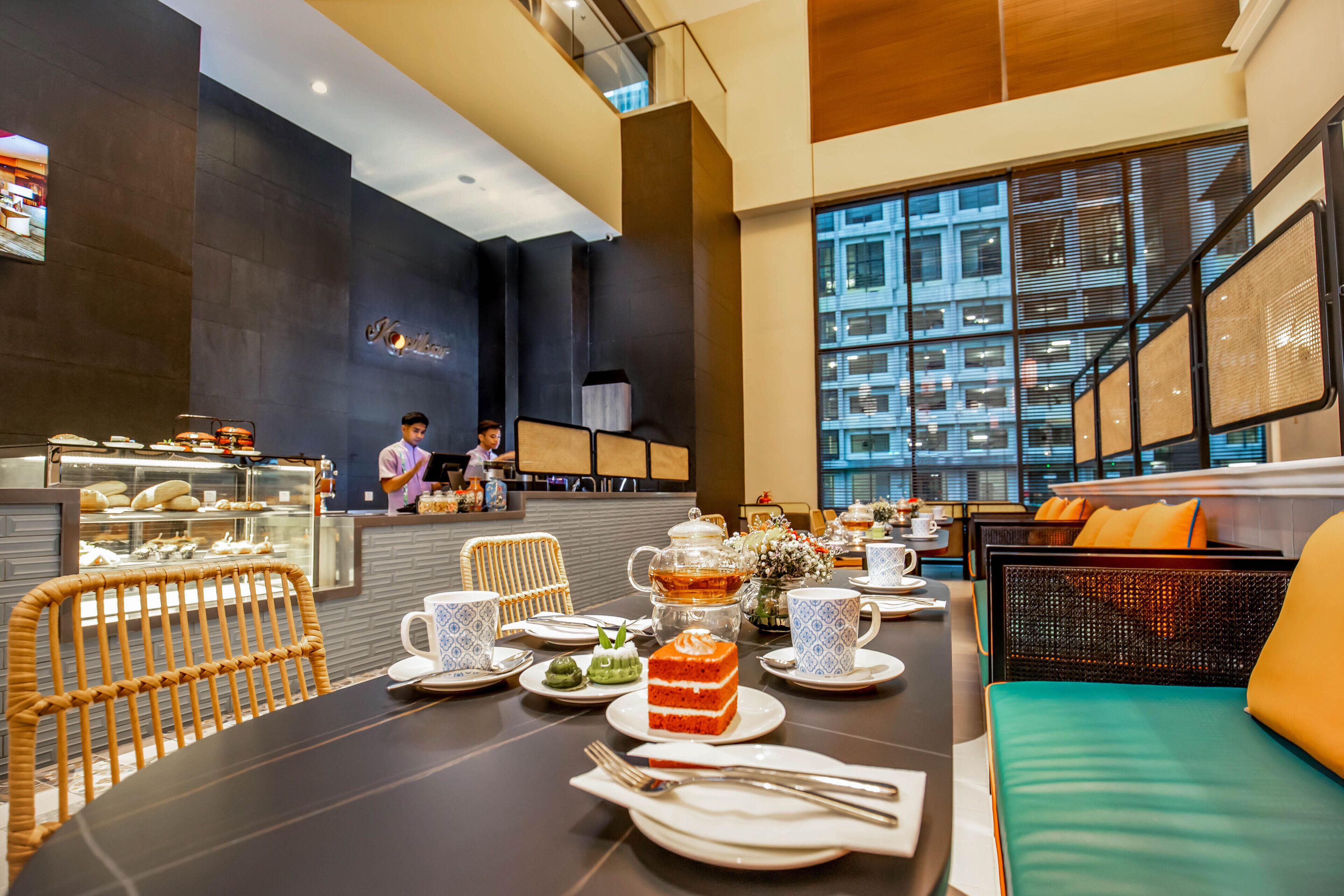A modern cafe with a long table set for tea service, wicker chairs, and a view of the city through large windows.
