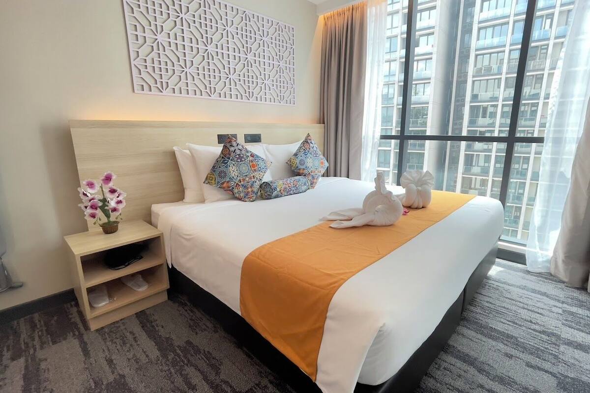 A hotel room with a large bed, a decorative wall panel, a window with curtains, and a small side table with a vase of flowers.