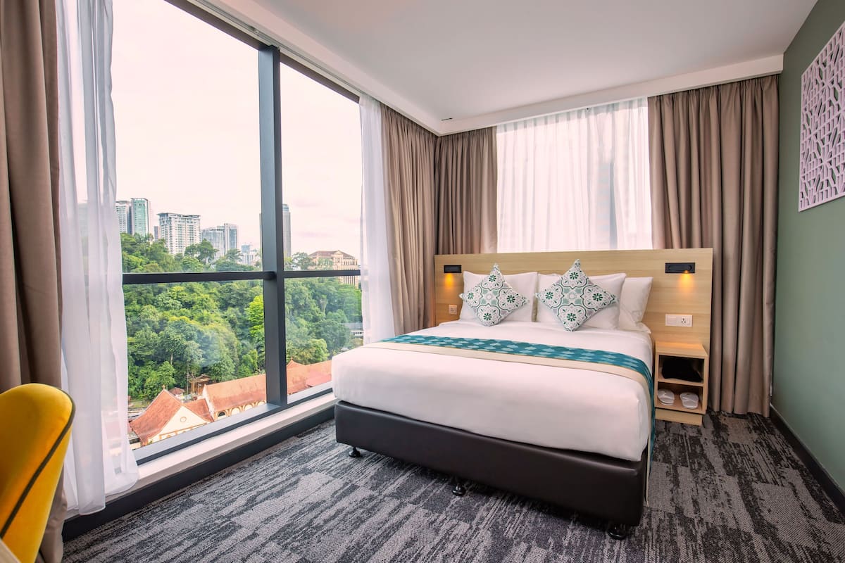 A hotel room with a large window, a bed with a wooden headboard, a bedside table, and a chair.