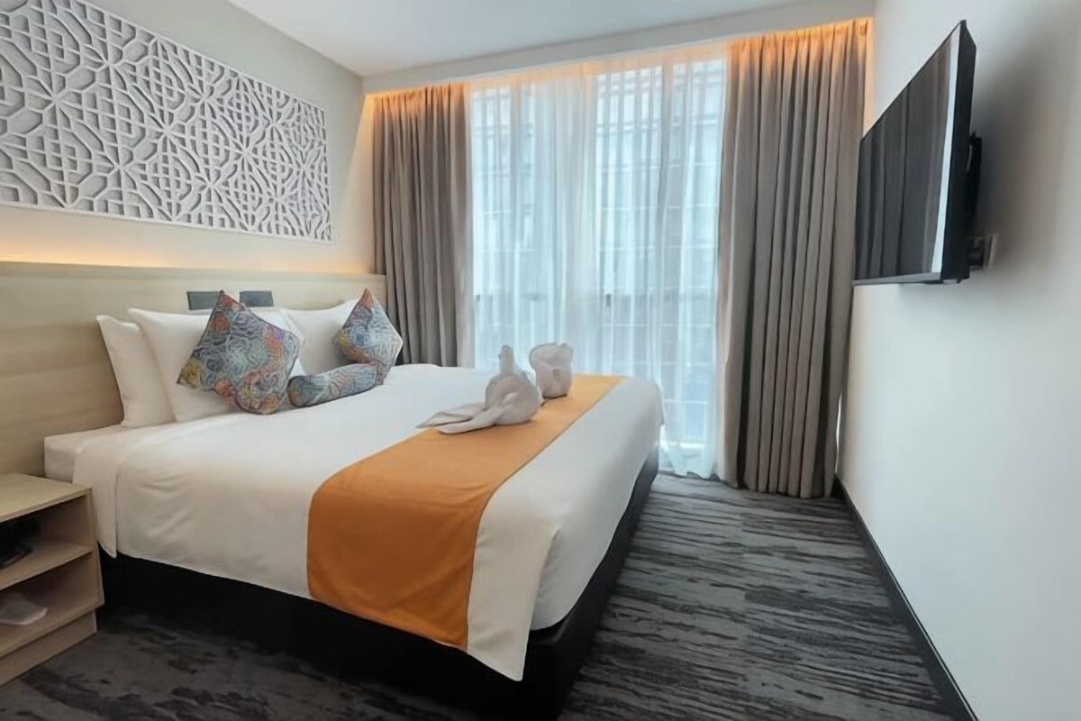 A hotel room with a large bed, a flat-screen TV, a decorative wall panel, and a window with curtains.