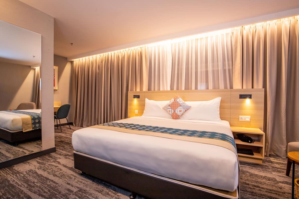 A hotel room with a large bed, a desk, a chair, a bedside table, and a mirror.