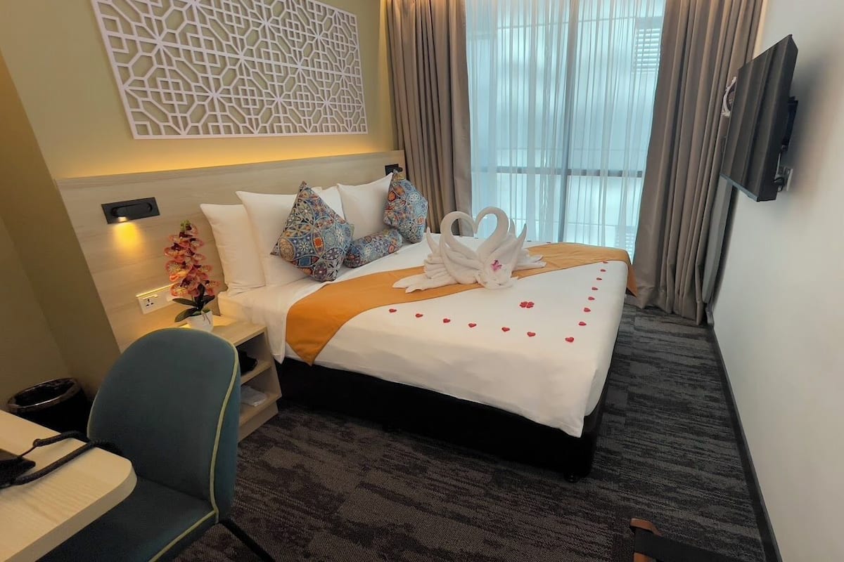 A hotel room with a neatly made bed, a TV mounted on the wall, and a window with curtains.