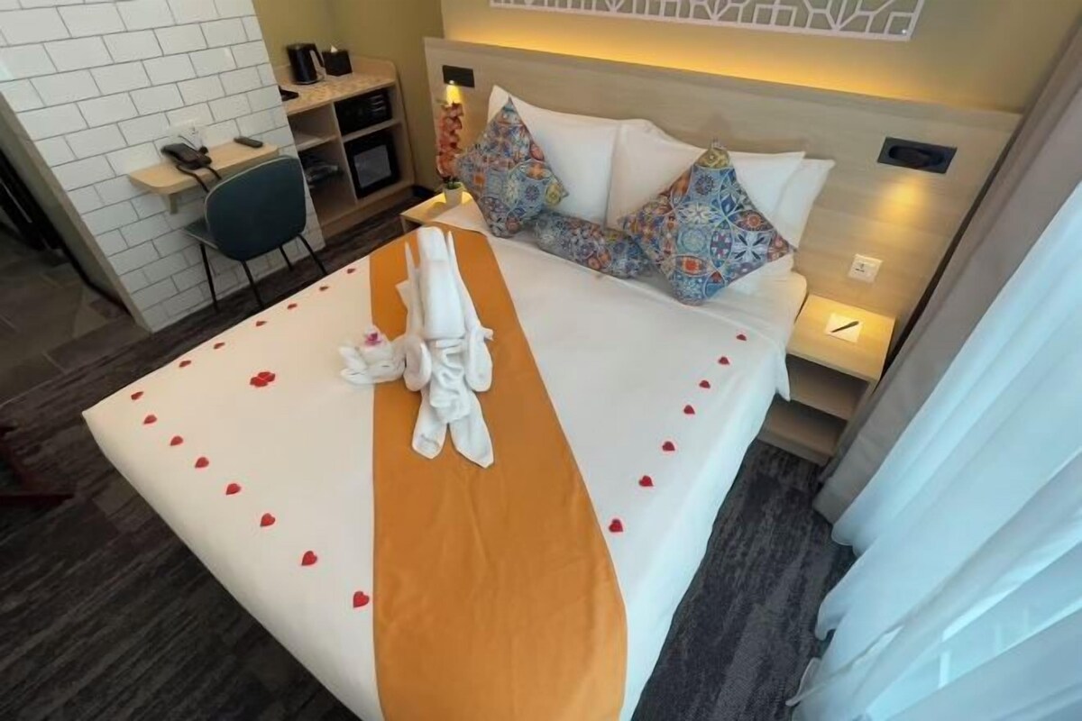 A hotel room with a neatly made bed, a small desk with a chair, and a bedside table with a lamp.