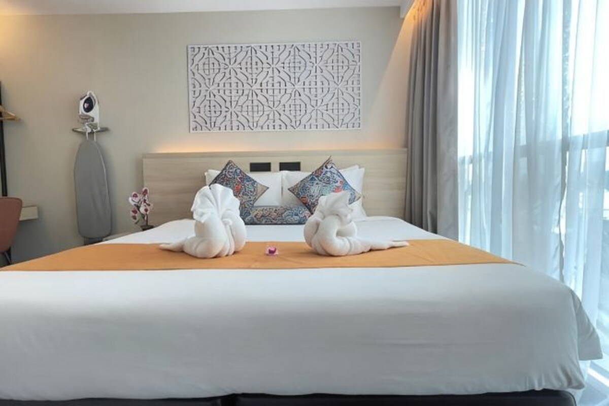 A hotel room with a bed, decorative pillows, a wall art piece, and a window with curtains.
