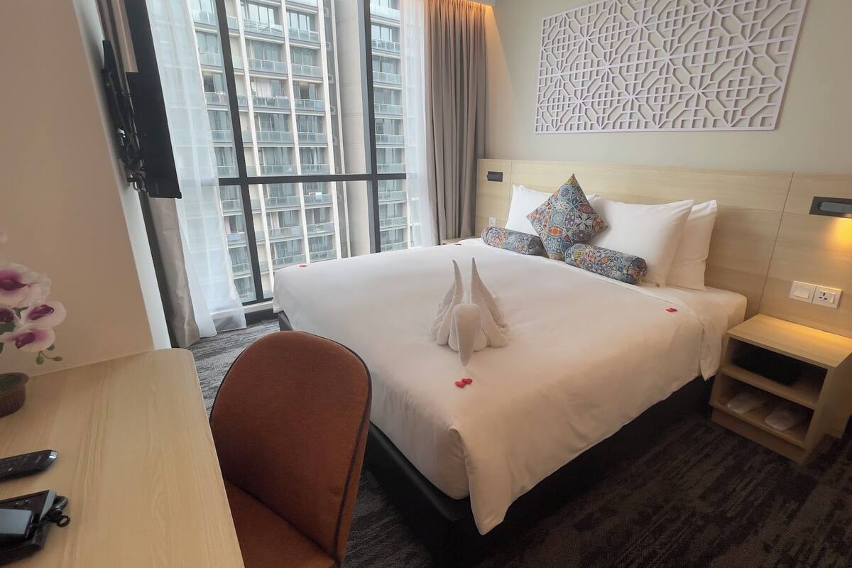 A hotel room with a large bed, a desk with a chair, a potted plant, and a view of a building through the window.
