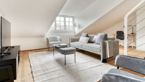 Quarto moderno no estilo loft com teto inclinado, uma janela grande, um sofá cinza, uma mesa de centro e uma TV em um suporte preto.