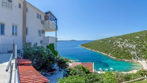 A coastal building with balconies overlooking a turquoise bay with boats and a hilly landscape.