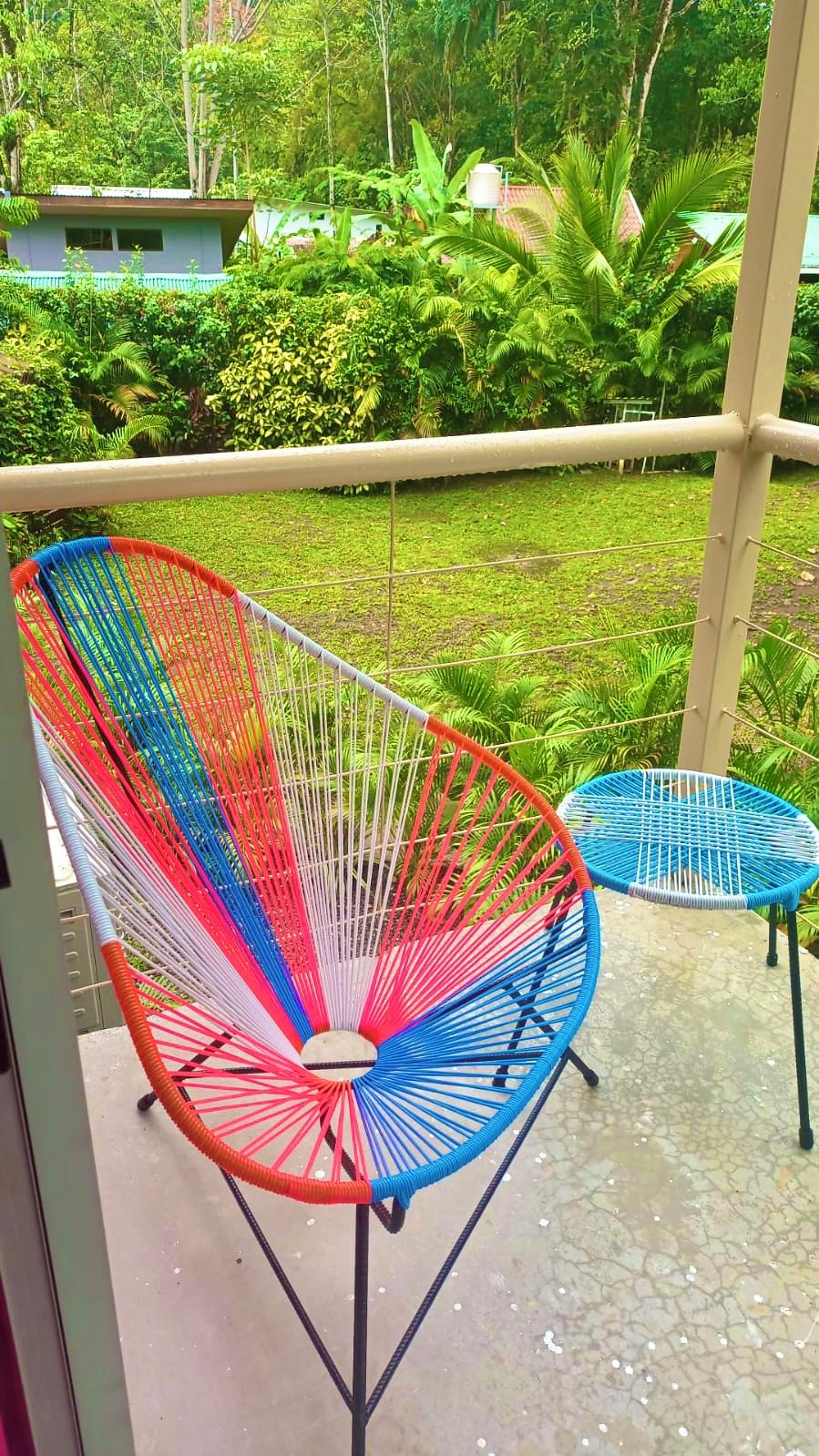 Un balcón con dos sillas tejidas de colores y una mesita pequeña, rodeado de exuberante follaje y un suelo de baldosas.