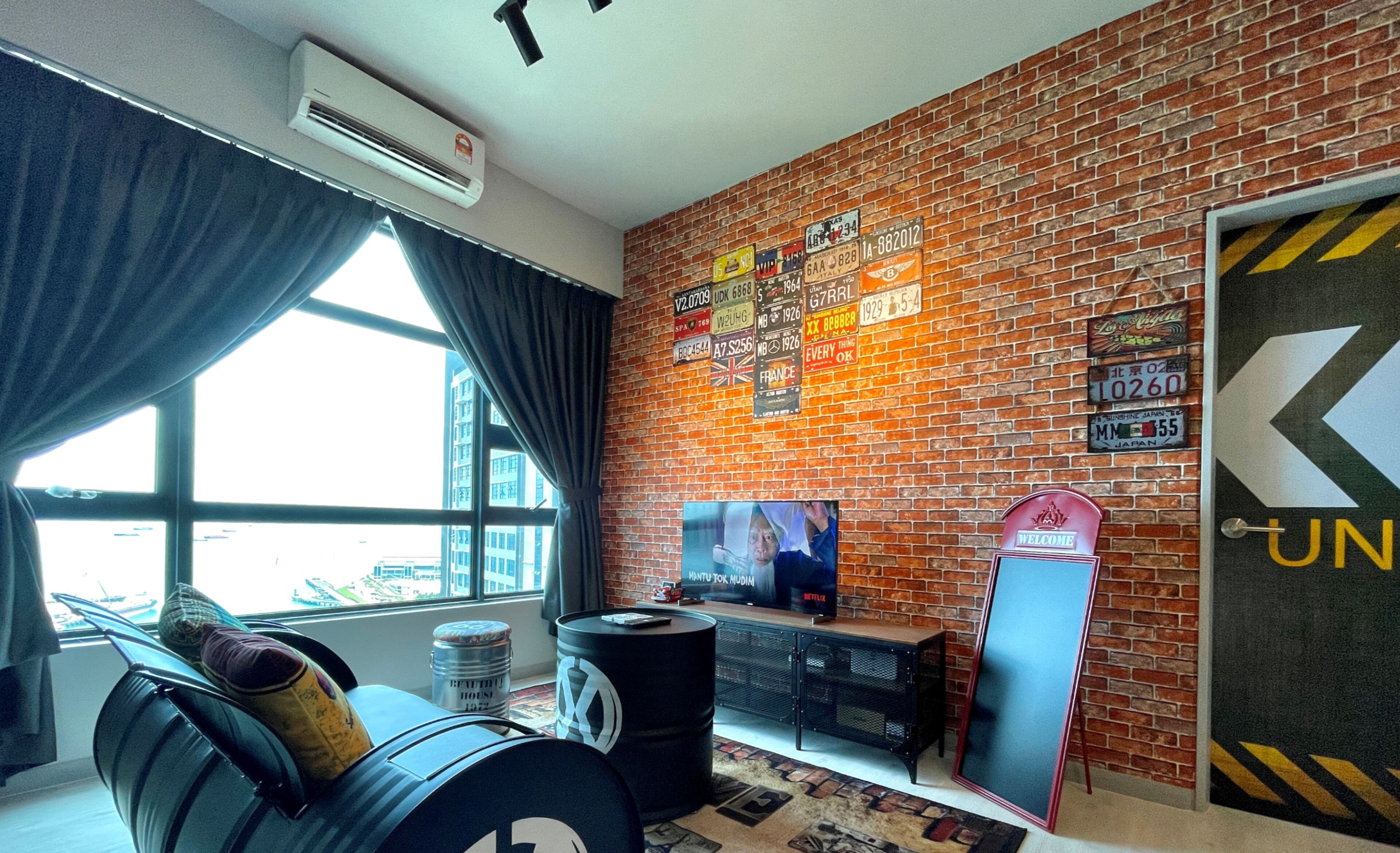 A room with a brick wall, a large window, a TV, and a 'Welcome' sign.