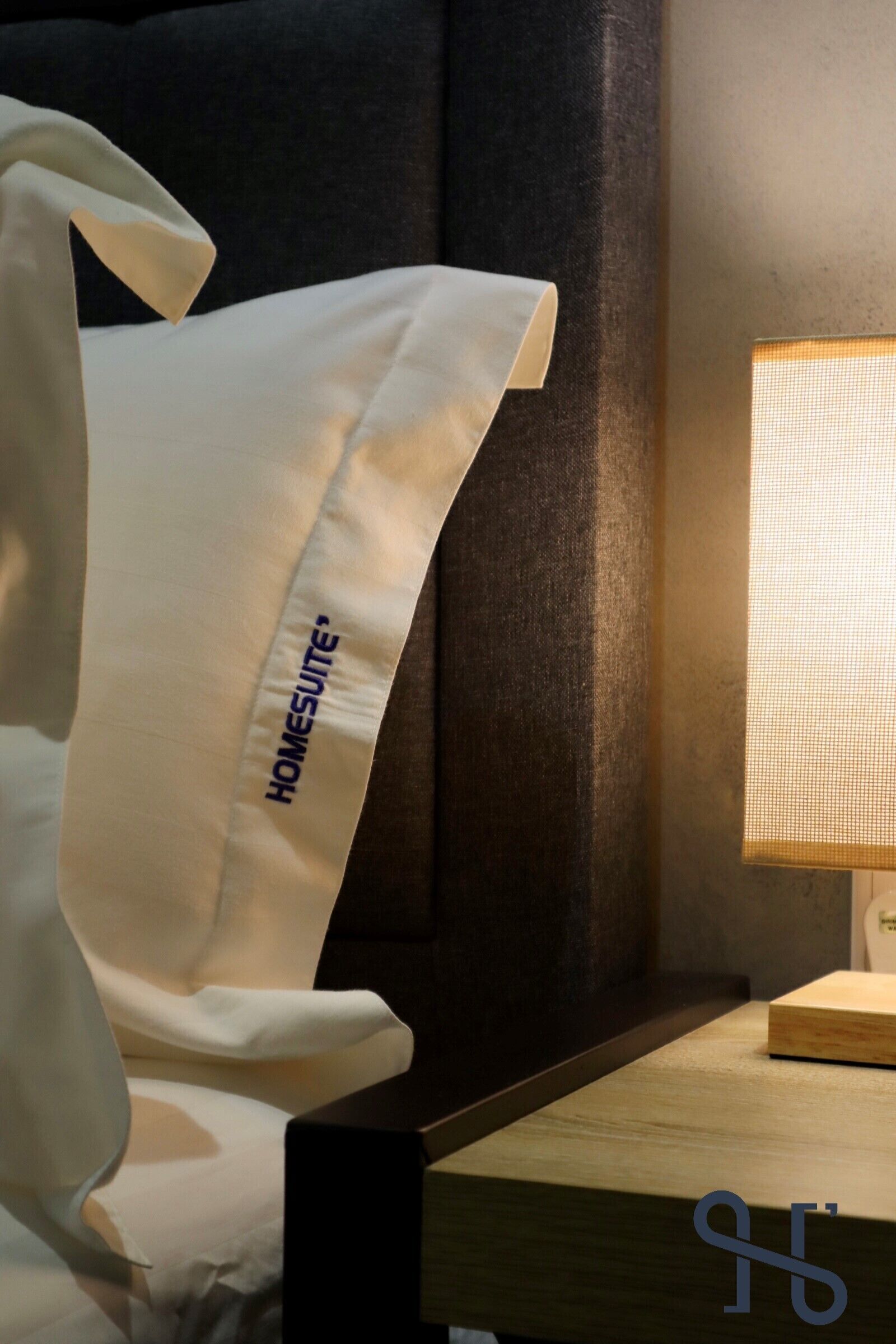Almohada blanca con el texto "HOMESUITE", una mesita de noche de madera y una lámpara con pantalla texturizada.