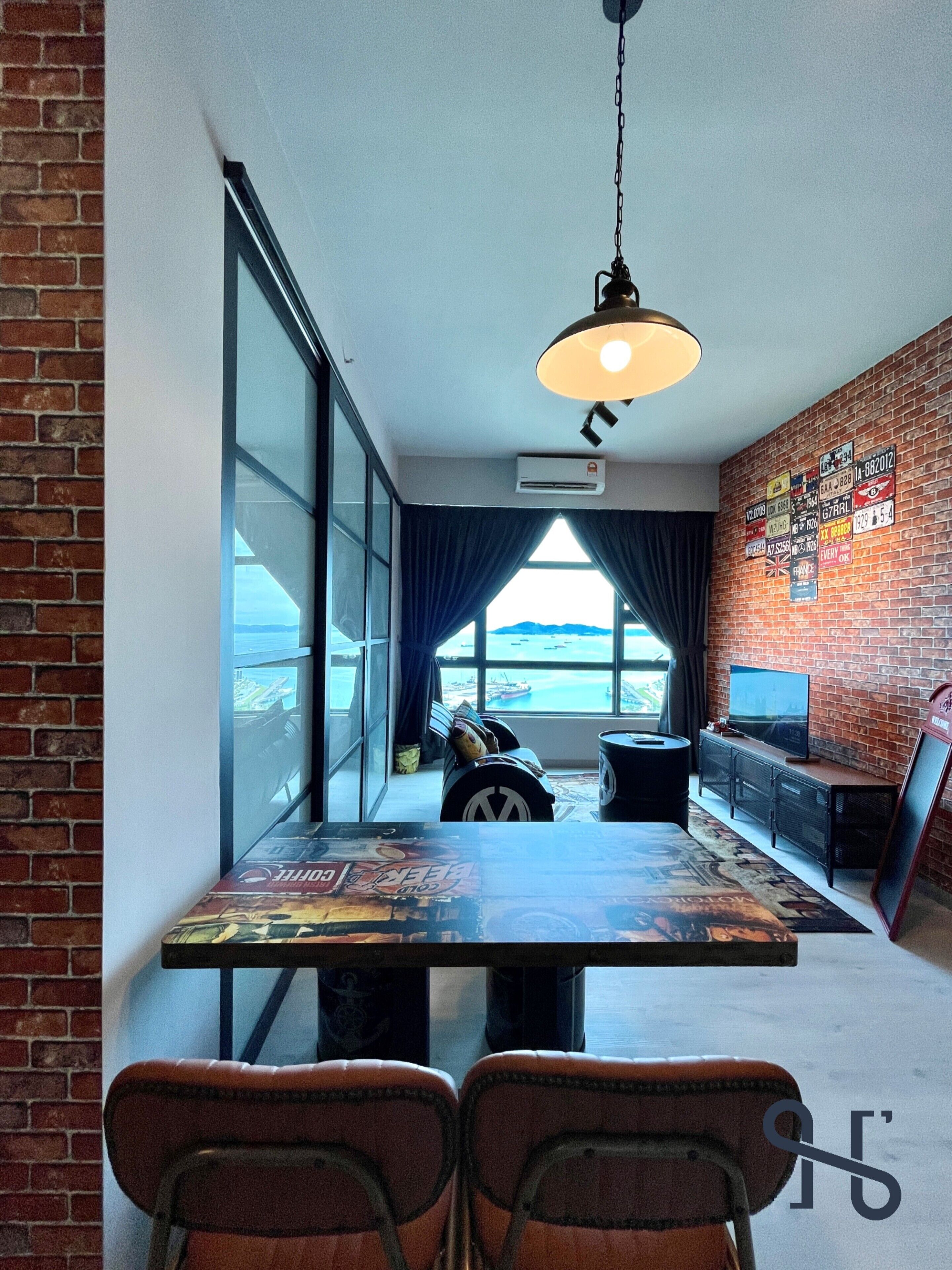 Une pièce moderne avec de grandes fenêtres, un mur en briques, une table avec des chaises, un luminaire suspendu et un téléviseur posé sur une étagère.