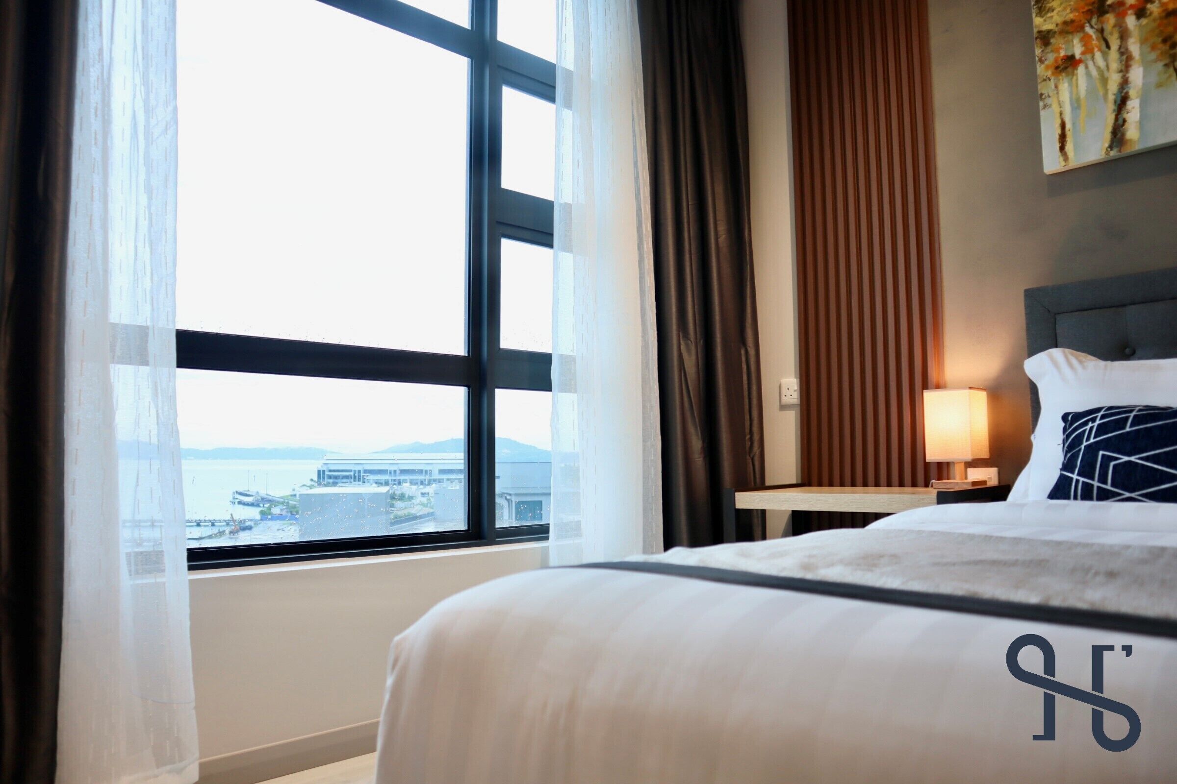 A hotel room with a large window, a bed with white bedding, a nightstand with a lamp, and a painting on the wall.