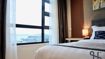 A hotel room with a large window, a bed with white bedding, a nightstand with a lamp, and a painting on the wall.
