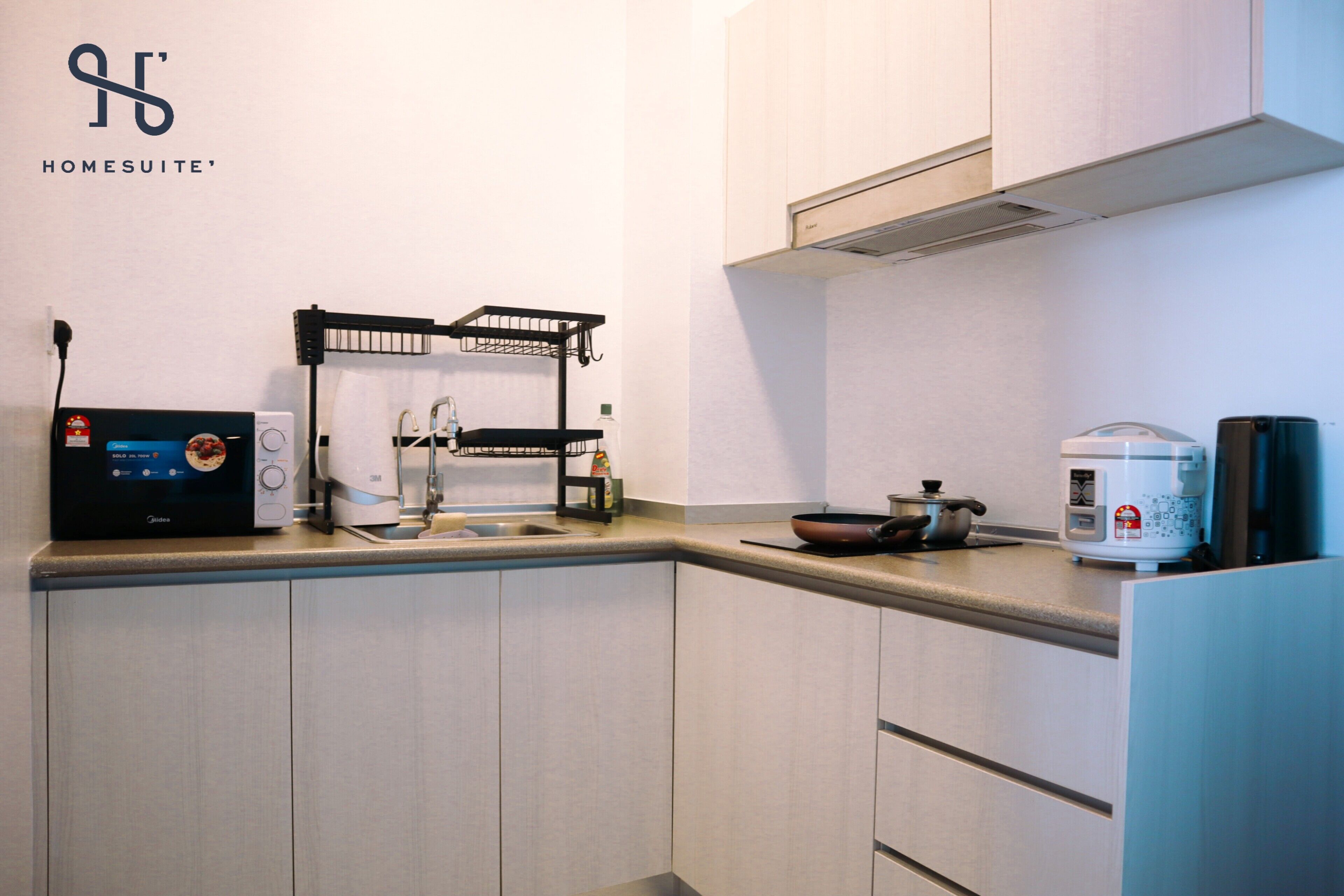 A modern kitchen with a microwave, kettle, and rice cooker on the counter, and a black drying rack on the wall.