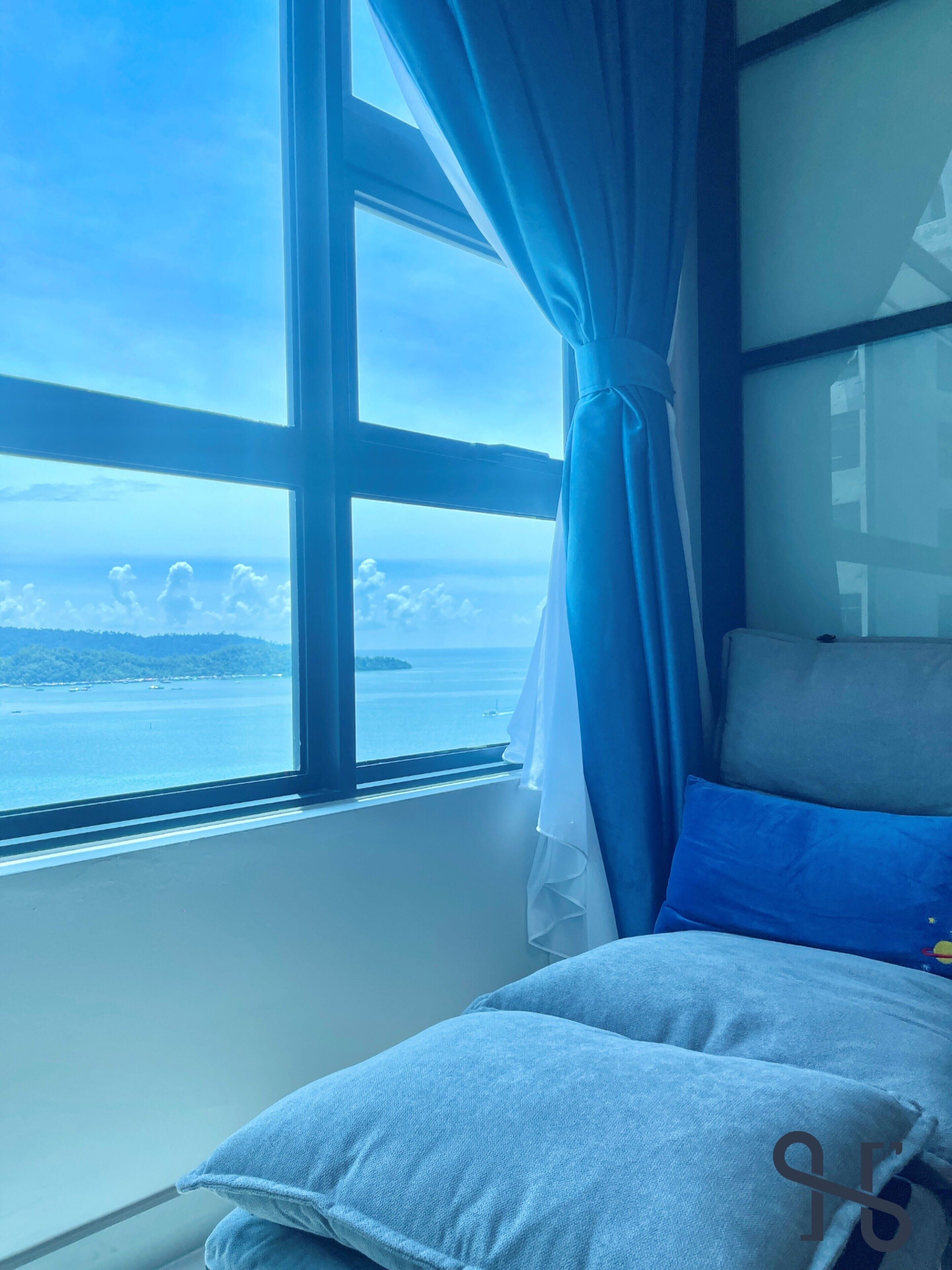 A room with a large window, a bed with blue and grey bedding, and a view of the sea and clouds.