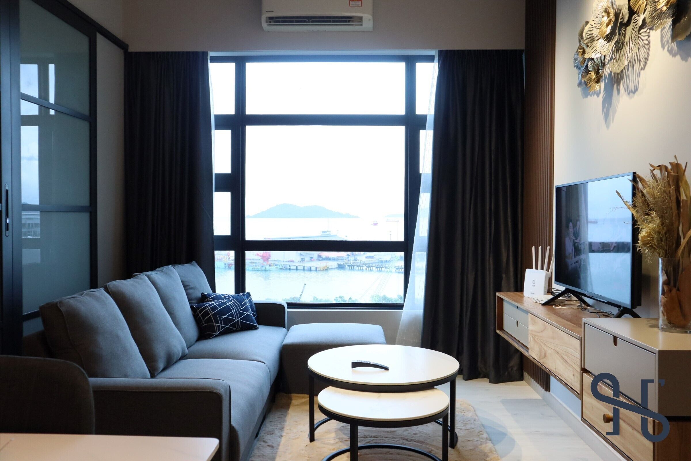 A modern living room with a sofa, a small table, a TV, and a view of a body of water through large windows.