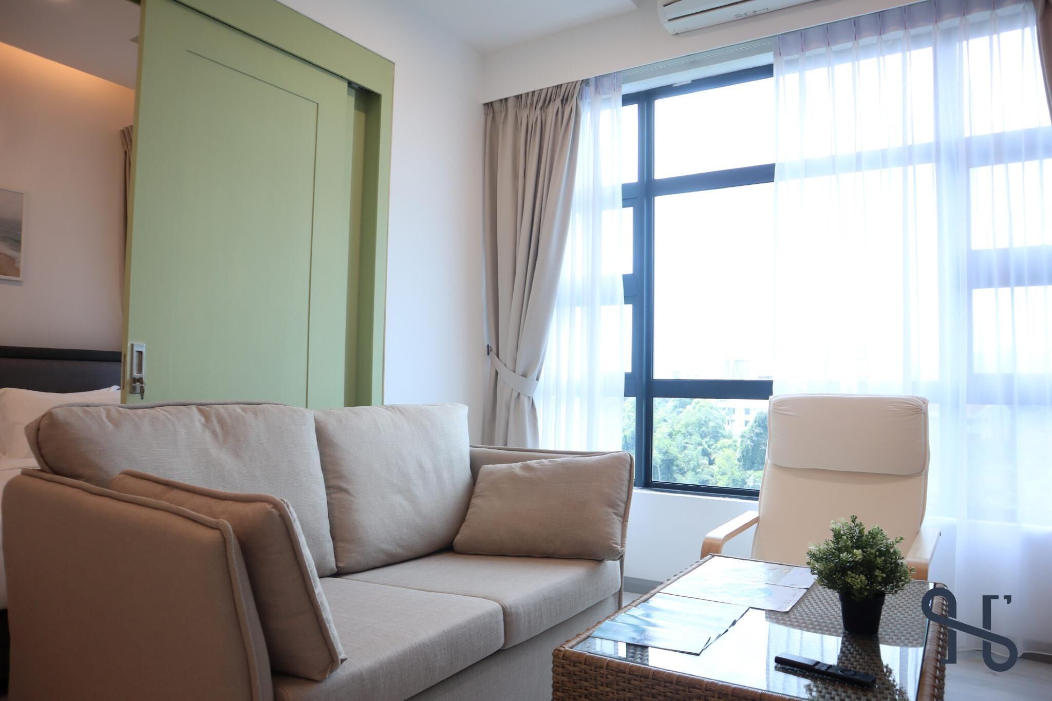 A modern hotel room with a beige sofa, a glass coffee table, a white armchair, and a large window with curtains.