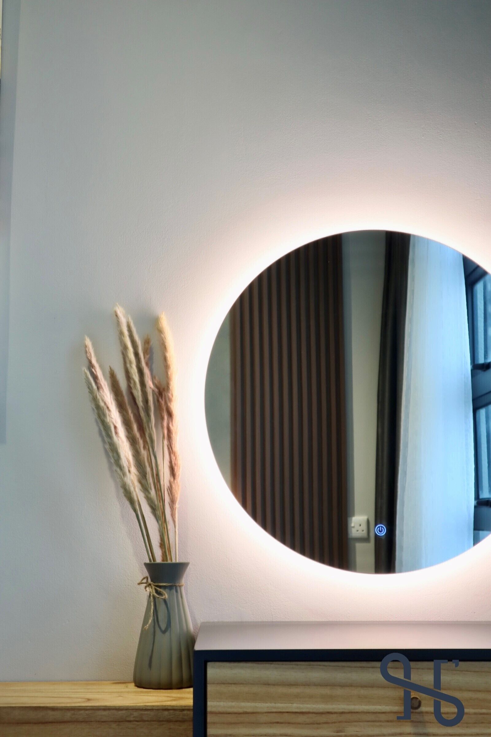 A round mirror with a lighted frame on a wooden surface, a vase with dried plants, and a plain wall.
